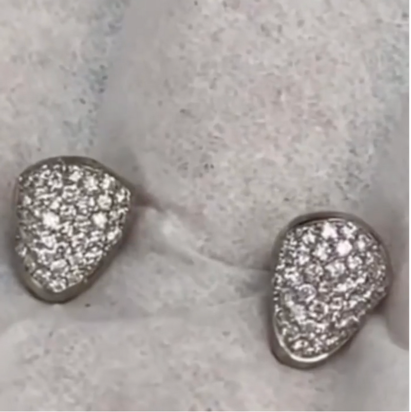 A pair of silver-toned teardrop-shaped stud earrings covered in small clear pavé stones, displayed on a textured light gray surface.